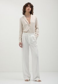 Donna con capelli ricci che indossa una camicia di seta beige, pantaloni bianchi a vita alta e ampi, scarpe open-toe e collana in catena argentata su uno sfondo neutro.