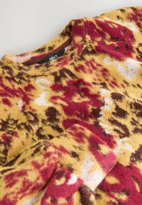 Fleecová mikina v žlto-červeno-hnedom abstraktnom vzore; dizajn s okrúhlym výstrihom; mäkká textúra; viditeľné švy a štítok.