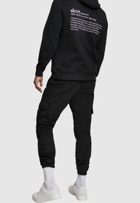 Felpa nera con cappuccio e scritta bianca sul retro, abbinata a pantaloni cargo neri. Il modello indossa sneakers bianche e calzini bianchi a altezza polpaccio.