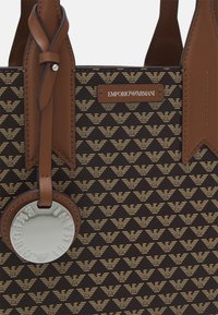 Hnědá kožená kabelka ve tvaru tote se geometrickým zlato vzorem loga, kovovým štítkem a dvěma horními uchy s akcentovanými detaily poblíž otvoru.