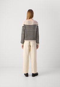 Stripet strikkegenser i beige og svart, med bred utringning og ribbede mansjetter. Kombinert med kremfargede vide bukser og svarte sko.