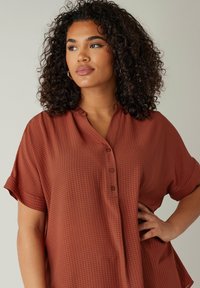 Camisa de manga corta de color óxido, texturizada con un patrón de cuadrícula, que presenta una tapeta con botones y un corte relajado. Tejido suave y ligero.