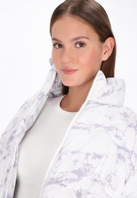 Padded jacket with a white and lavender marble pattern, featuring a high collar and zipper. Worn over a white long-sleeve shirt.