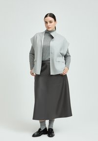 Graue ärmellose Weste über einem grauen Rollkragenpullover, kombiniert mit einem langen, dunkelgrauen Lederminirock und schwarzen Slippern, mit sichtbaren grauen Socken.