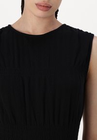 Melna bez piedurknēm kleita ar apaļu izgriezumu, kurai ir tekstūras audums un samontēta detaļa viduklī.