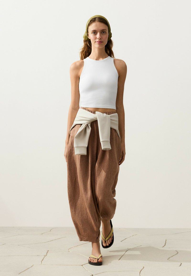 Jeune femme aux cheveux ondulés portant un crop top blanc sans manches, un pantalon large marron, un sweat-shirt noué autour de la taille et des tongs, marchant sur un sol carrelé.