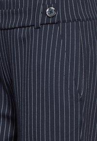 Pantalons rayés bleu marine en tissu lisse, avec des rayures verticales blanches, une fermeture éclair, des poches latérales et une fermeture à bouton unique.