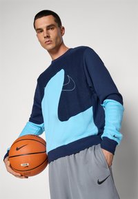 Sweatshirt da Nike com uma combinação de materiais azul-marinho e azul claro, designs texturizados e um logótipo em destaque. Combinado com calções cinzentos e uma bola de basquetebol.
