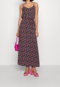 Robe maxi florale avec bretelles spaghetti réglables, fond noir, motif de fleurs roses et blanches, taille cintrée, assortie à des talons compensés roses et un sac.