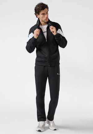 Giacca nera con zip e dettagli bianchi, abbinata a pantaloni della tuta neri. Presenta una texture morbida e un design sportivo. Sneakers argento completano l'outfit.