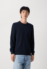Marineblauer Strickpullover mit rundem Halsausschnitt und gerippten Bündchen, kombiniert mit hellblauen Jeans. Strukturmuster auf dem Pullover sichtbar.