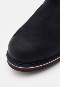 Musta mokkanahkainen nilkkuri pyöreällä kärjellä, jossa on kontrastivärinen laivastonsininen pohja ja punavalkoiset raidat sivulla. Pinnan tekstuuri.