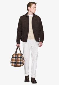 Brun ruskindsjakke med to frontlommer, beige strikket sweater, hvide bukser og en rund plaid taske i brun, sort og cream. Brune sko.