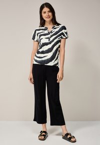 Zwart-witte t-shirt met zebra-strepen en knopen bij de halslijn, gecombineerd met zwarte wijde broek en zwarte sandalen met een strikdetail.
