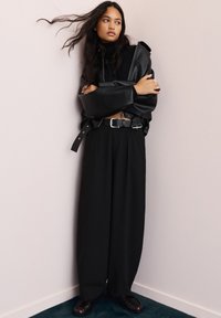 Črna usnjena prenaglašena jakna z širokimi rokavi, v kombinaciji z visokim črnim hlačami in črnimi čevlji do gležnjev. Preprost, minimalističen dizajn.