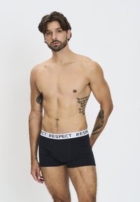 Fekete pamut boxernadrág fehér "RESPECT" derékszíjjal, amelyek szoros illeszkedésűek és sima tapintásúak. A modelltetoválások láthatók a karján.