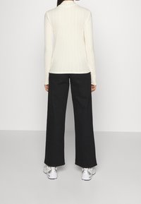 Vaalean beige ribatti pitkähiha klubi, jota on yhdistetty mustiin leveisiin housuihin. Jalkineissa on metalliset yksityiskohdat. Minimalistinen muotoilu yksinkertaisen taustan vasten.