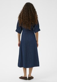 Denim maksi obleka v temno modri barvi, ki ima kratke napihnjene rokave in prilegajoč trup, ter razširjen krilo, ki sega do sredine meč. Oblečene rjave ravne čevlje.