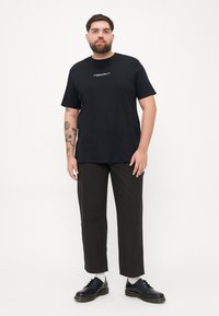 Black cotton t-shirt with short sleeves and centered white text, paired with dark pants and black lace-up shoes. Simple, casual design.