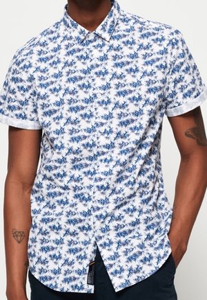 Weißes Hemd mit kurzen Ärmeln und blauer Palmenmusterung, getragen von einer Person mit Tätowierungen an beiden Armen.