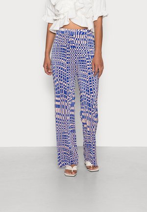 Unterkörper einer Person, die hochtaillierte, weit geschnittene Hosen mit einem rosa-blauen verzerrten Schachbrettmuster und weißen offenen Sandalen trägt.