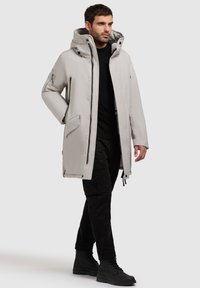 Jachetă parka gri deschis cu guler înalt, glugă supradimensionată și închidere cu button în față. Are buzunare laterale și o textură elegantă, netedă.