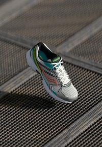 Scarpa da corsa Saucony bianca e turchese con dettagli arancioni, posata su una superficie di grata metallica testurizzata al sole.