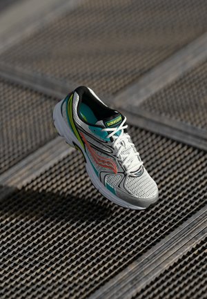 White and teal Saucony running shoe with orange accents resting on a textured metal grate surface in sunlight.