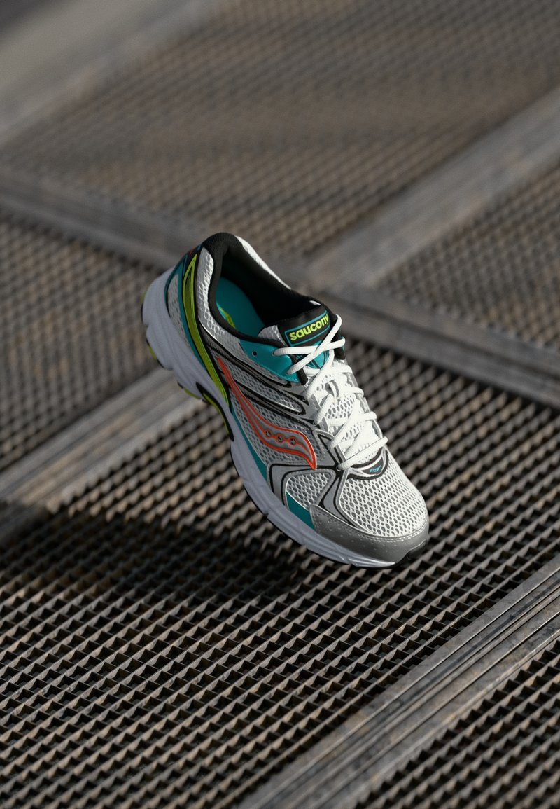 Scarpa da corsa Saucony bianca e turchese con dettagli arancioni, posata su una superficie di grata metallica testurizzata al sole.