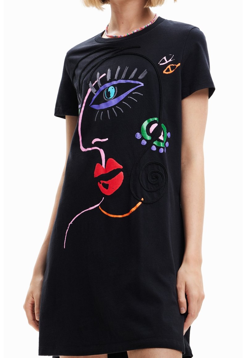 Robe noire avec un motif abstrait coloré représentant un visage avec un œil bleu, des lèvres rouges et une boucle d'oreille verte, portée par une personne aux cheveux blonds courts.