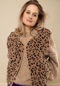 Donkerbruine fuzzy vest met zwart luipaardprint, afgewerkt met beige. Gedragen over een crèmekleurige gebreide trui met een ronde halslijn en een gestructureerde stof.