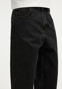 Gros plan d'une personne portant un jean en denim noir et une chemise blanche, montrant la poche avant et les détails des coutures.