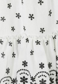 Tissu blanc avec broderie florale noire et motif festonné. Volant froncé à l'ourlet, ajoutant texture et détail.