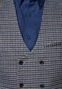 Dwurzędowa kamizelka w wzór houndstooth, z szaro-czarnymi i niebieskimi tonami. Teksturowana tkanina z czarnymi guzikami i niebieską koszulą pod spodem.