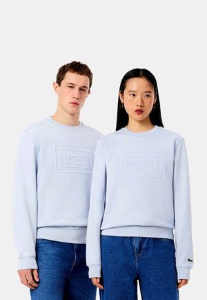 To unge voksne iført lyseblå Lacoste sweatshirts og blå jeans står side om side mod en ensfarvet baggrund.