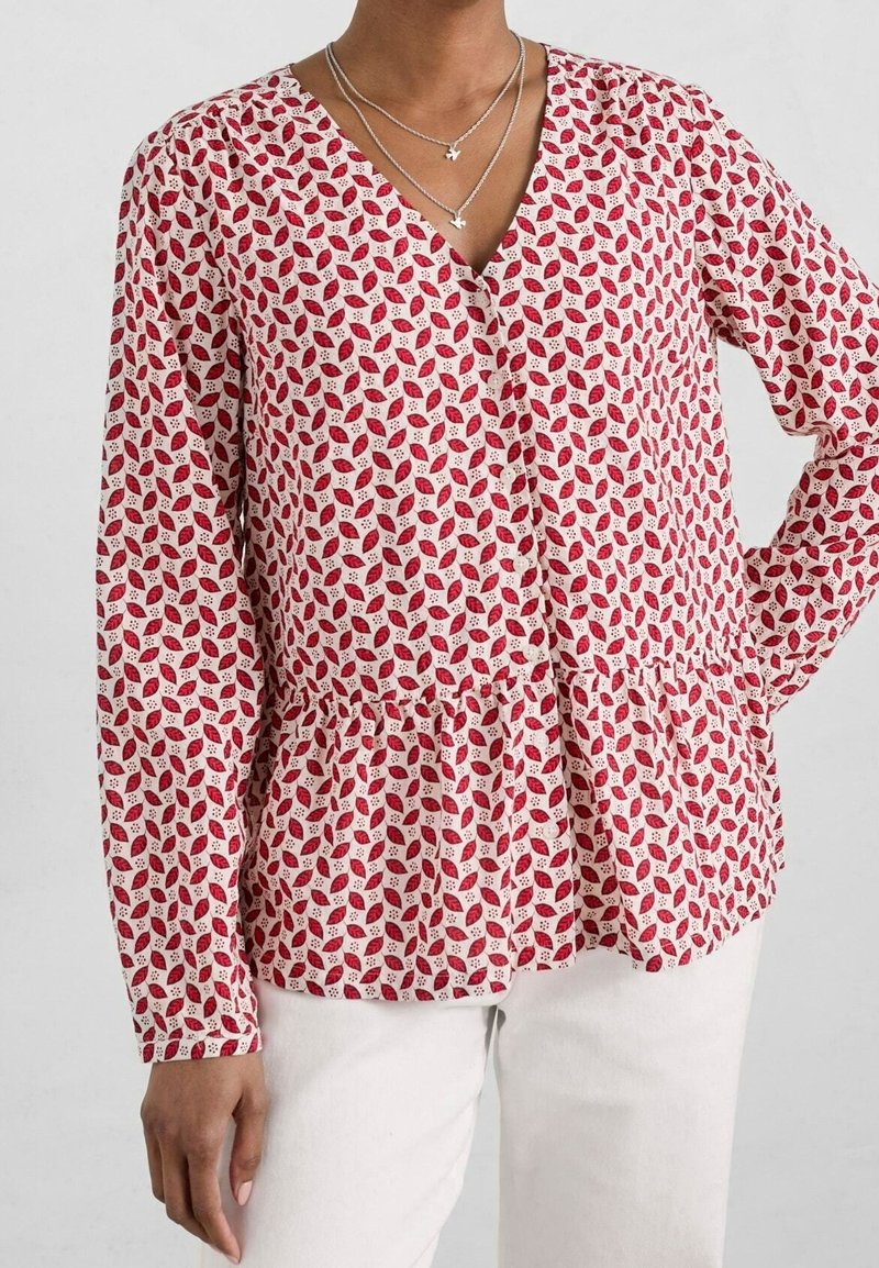 Femme portant un chemisier à manches longues avec un motif de feuilles rouges et blanches et un col en V, associé à un pantalon blanc et des colliers en argent superposés.