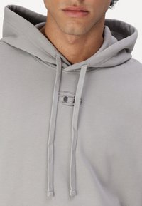 Hellgraue Kapuzenjacke mit texturiertem Finish, ausgestattet mit einer Kordelzugkapuze und einem silbernen ovalen Logodetail auf der Brust.