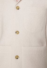 Kremowa wełniana kurtka o fakturowanej powierzchni, z wyciętym kołnierzem i dwoma przednimi kieszeniami. Detale tworzą mosiężne guziki.