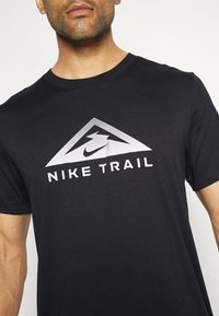 Hombre vistiendo una camiseta negra de Nike Trail con un logo triangular plateado y texto centrado en el pecho.