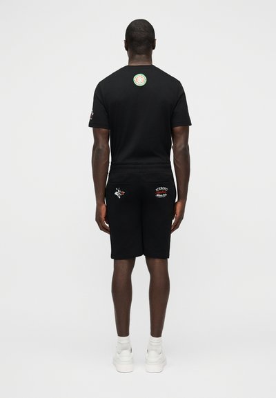 Homme debout de dos portant un t-shirt noir et un short avec de petites patches colorées, des chaussettes blanches et des baskets blanches sur un fond uni.