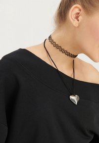 Musta olkapäät paljastava collegepaita, jossa on resoroitu kaulus, yhdistetty mustaan choker-kaulakoruun ja hopeiseen syd�änriipukseen mustalla nauhalla.