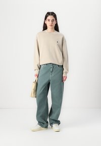 Felpa beige con logo, pantaloni mint green larghi, sneakers bianche e una piccola borsa beige in mano. Outfit semplice e casual.
