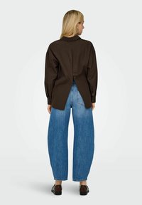 Hnědá oversized košile s knoflíkovou aplikací na zádech, spárovaná s modrými džíny s vysokým pasem a širokými nohavicemi a černými botami.