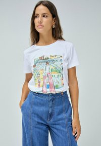 Camiseta de algodón blanca con un diseño impreso colorido que presenta una figura con pantalones a rayas y un fondo de paisaje urbano. Jeans de mezclilla de cintura alta.