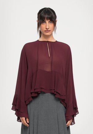 Bordeaux sheer bluse med lagdelte volanger og en nøkkelhullsutringning. Lange ermer med utsvingte kanter. Brukt over et grått plisserte skjørt.