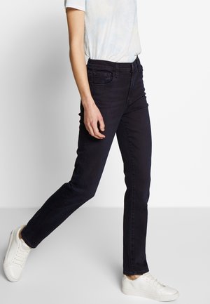 Person, die schwarze enge Jeans, weiße Turnschuhe und ein helles kurzärmeliges T-Shirt trägt, vor einem schlichten weißen Hintergrund läuft.