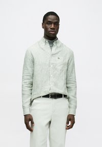 Zöld-fehér csíkos, gombos ing klasszikus gallérral, bal mellrészen zsebbel és apró logóval a mellkason. Világos nadrággal viselve.