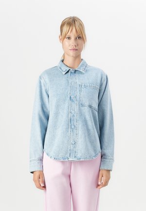 Jeune femme portant une chemise en denim clair à boutons avec une poche sur la poitrine et un pantalon rose clair, se tenant devant un fond blanc.