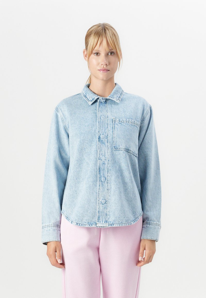 Jeune femme portant une chemise en denim clair à boutons avec une poche sur la poitrine et un pantalon rose clair, se tenant devant un fond blanc.