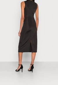 Schwarzes ärmelloses Midi-Kleid mit enger Silhouette, einem Rückenschlitz und glatter Textur. Kombiniert mit schwarzen Knöchelriemen-Absätzen.